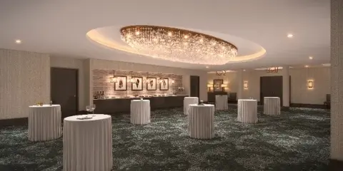 A spacious event ballroom featuring a large circular chandelier and multiple white-draped high-top tables.