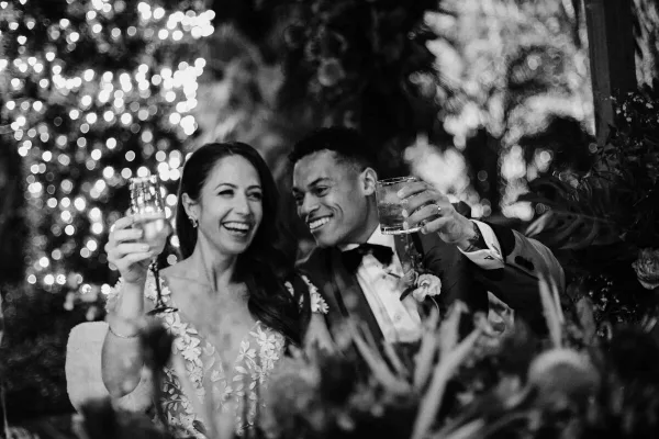 A black and white photograph of a smiling bride and groom toasting with champagne glasses at a wedding reception.