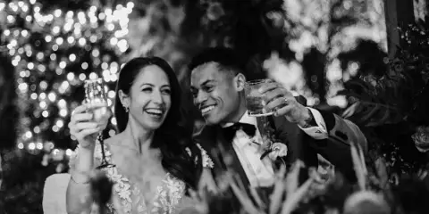 A black and white photograph of a smiling bride and groom toasting with champagne glasses at a wedding reception.