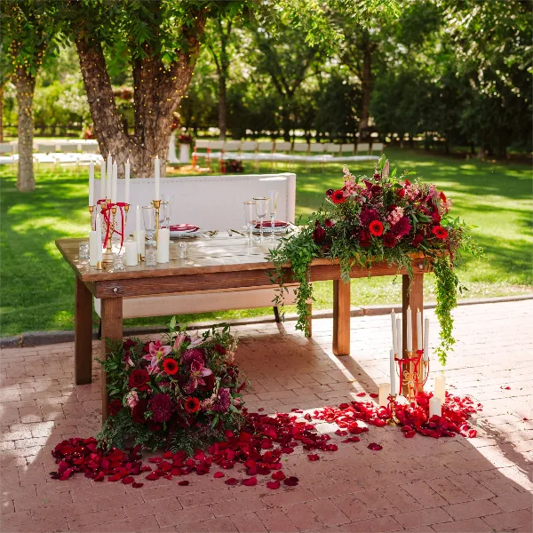 A wooden wedding table set with a white bench, candles, and red floral arrangements in an outdoor garden setting.