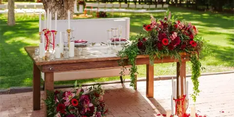 A wooden wedding table set with a white bench, candles, and red floral arrangements in an outdoor garden setting.