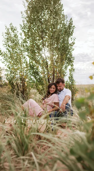 A couple sitting together in a grassy field with trees in the background, overlaid with the text 'Life, Unscripted.'