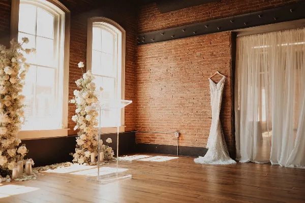 Interior of a historic brick venue with a wedding dress hanging on the wall and floral arrangements near arched windows.