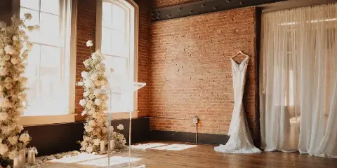 Interior of a historic brick venue with a wedding dress hanging on the wall and floral arrangements near arched windows.