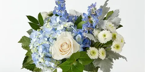 A floral arrangement featuring blue delphiniums, light blue hydrangeas, a white rose, and white chrysanthemums in a clear glass vase.