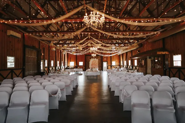Interior of a rustic barn wedding venue with rows of white chairs, chandeliers, and draped fabric.