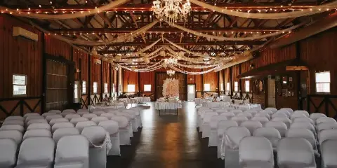 Interior of a rustic barn wedding venue with rows of white chairs, chandeliers, and draped fabric.
