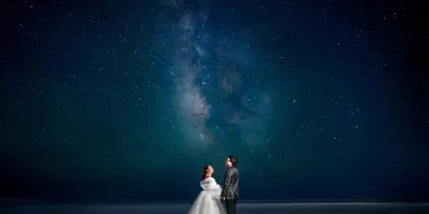 A bride and groom standing together under a starry night sky with the Milky Way visible above them.