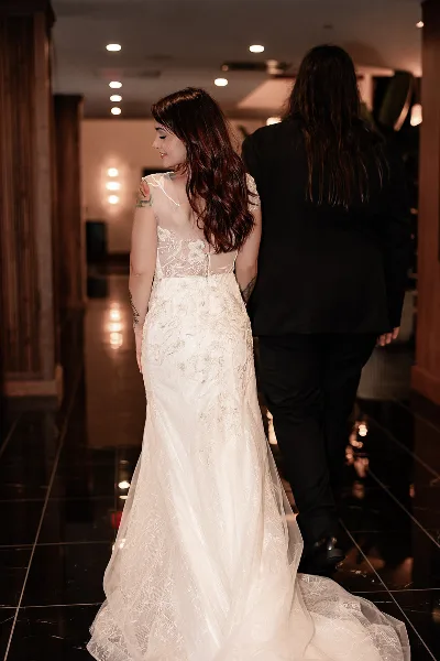 A bride in a lace wedding gown walking arm-in-arm with a partner in a dark suit through a warmly lit venue hallway.