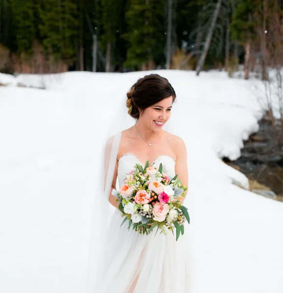 A smiling bride in a white wedding dress and veil holding a bouquet of flowers in a snowy outdoor setting.
