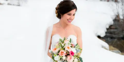 A smiling bride in a white wedding dress and veil holding a bouquet of flowers in a snowy outdoor setting.