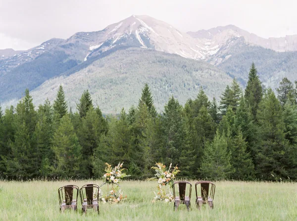 Outdoor wedding ceremony setup with floral arrangements and chairs in a grassy field with mountains in the background.