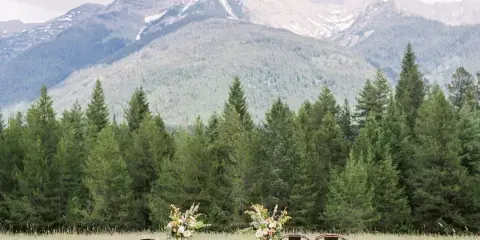Outdoor wedding ceremony setup with floral arrangements and chairs in a grassy field with mountains in the background.