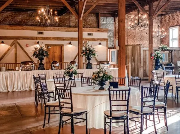 Interior of a historic event venue with exposed wooden beams, brick walls, chandeliers, and round tables set for a wedding.