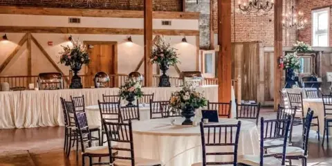 Interior of a historic event venue with exposed wooden beams, brick walls, chandeliers, and round tables set for a wedding.