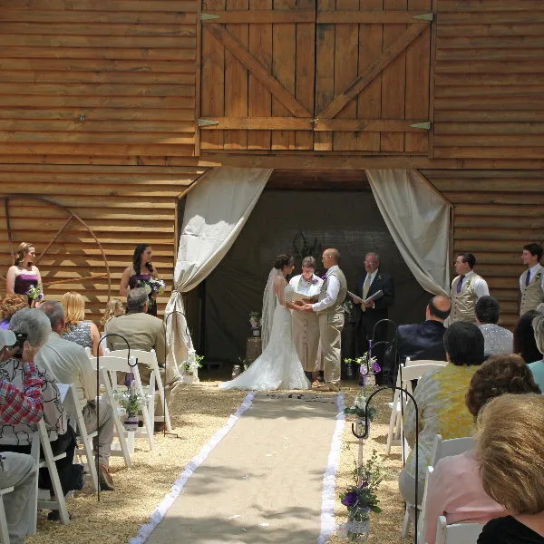 A wedding ceremony in progress at a wooden barn venue with a bride and groom at the altar and guests seated in white chairs.