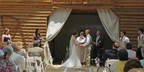 A wedding ceremony in progress at a wooden barn venue with a bride and groom at the altar and guests seated in white chairs.