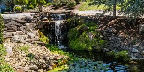 A scenic view of a wedding venue featuring a stone waterfall, a pond with lily pads, and a wooden gazebo in the background.