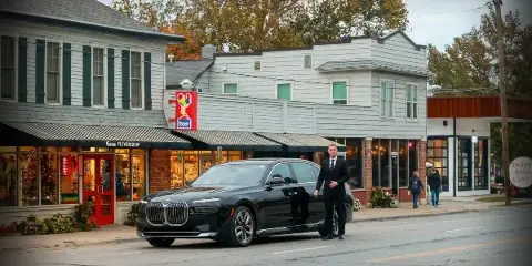 A professional chauffeur in a dark suit stands beside a black luxury sedan parked on a street in front of storefronts.