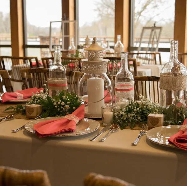 A rustic wedding table setting featuring glass bottle centerpieces, burlap accents, and candles.