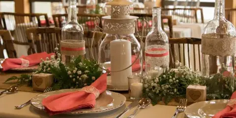 A rustic wedding table setting featuring glass bottle centerpieces, burlap accents, and candles.