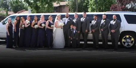 A wedding party including the bride, groom, and attendants posing in front of a white limousine.
