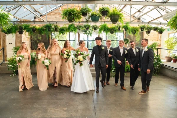 A wedding party consisting of a bride, groom, bridesmaids, and groomsmen walking through a greenhouse venue.