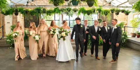 A wedding party consisting of a bride, groom, bridesmaids, and groomsmen walking through a greenhouse venue.