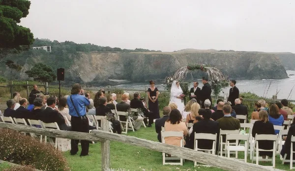 A wedding ceremony taking place outdoors with an officiant, bride, and groom standing before a floral arch, with seated guests and a coastal cliff backdrop.