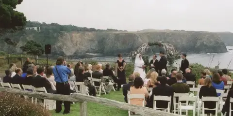 A wedding ceremony taking place outdoors with an officiant, bride, and groom standing before a floral arch, with seated guests and a coastal cliff backdrop.