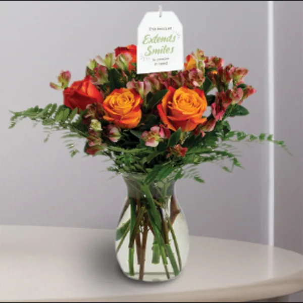 A floral arrangement featuring orange roses and pink snapdragons in a clear glass vase with a hanging tag.
