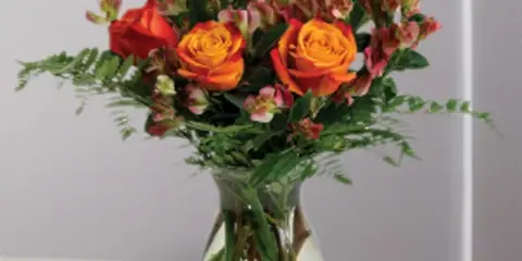 A floral arrangement featuring orange roses and pink snapdragons in a clear glass vase with a hanging tag.