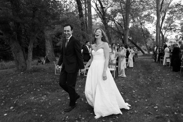 A black and white photograph of a bride and groom walking down an outdoor aisle, laughing, with wedding guests seated on either side.