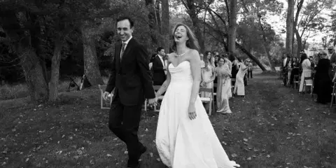 A black and white photograph of a bride and groom walking down an outdoor aisle, laughing, with wedding guests seated on either side.