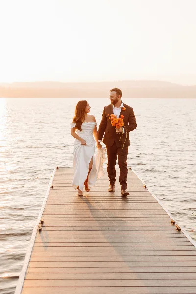 A bride and groom walking hand-in-hand on a wooden dock extending into a lake at sunset.