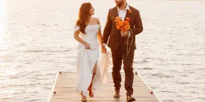 A bride and groom walking hand-in-hand on a wooden dock extending into a lake at sunset.