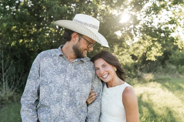 A smiling couple posing outdoors in a grassy field with trees in the background.