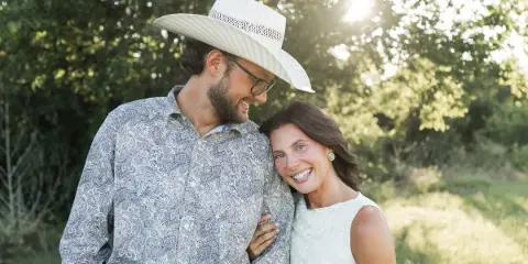 A smiling couple posing outdoors in a grassy field with trees in the background.