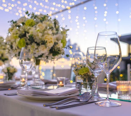 Elegant catering table setting with white plates, wine glasses, silverware, and floral arrangements under string lights.
