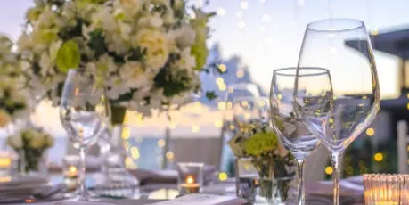 Elegant catering table setting with white plates, wine glasses, silverware, and floral arrangements under string lights.