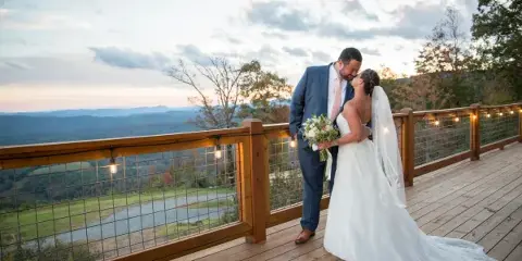 A bride and groom embracing on a wooden deck with a scenic mountain view in the background.