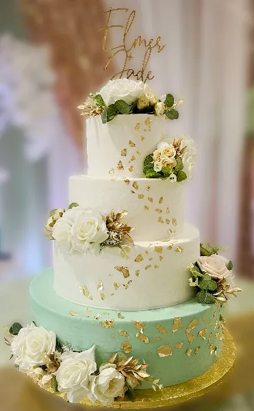 A four-tier wedding cake featuring white and mint green tiers, adorned with white roses, greenery, and gold leaf accents.