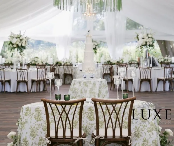 A wedding reception table setting featuring specialty wooden chairs and floral patterned table linens, with a LUXE Event Linen watermark.
