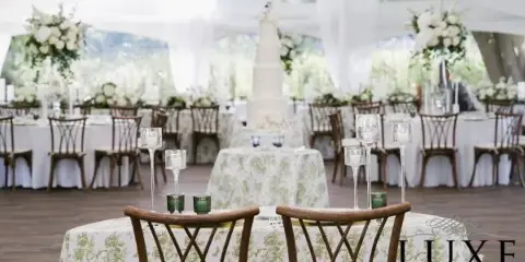A wedding reception table setting featuring specialty wooden chairs and floral patterned table linens, with a LUXE Event Linen watermark.
