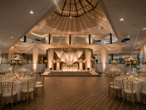 A spacious wedding reception hall with high ceilings, a decorated stage, and round tables set for a celebration.