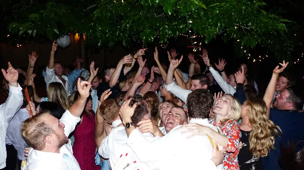 A crowded dance floor at a wedding reception with guests dancing and raising their hands under string lights.