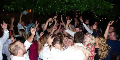 A crowded dance floor at a wedding reception with guests dancing and raising their hands under string lights.