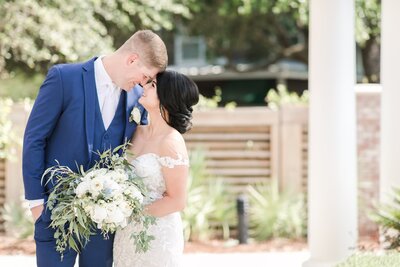 A bride and groom in wedding attire sharing a moment outdoors with a white bouquet.