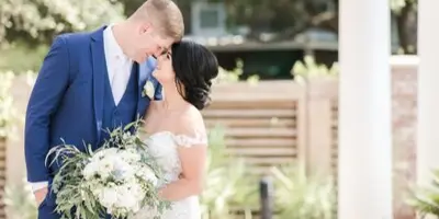 A bride and groom in wedding attire sharing a moment outdoors with a white bouquet.