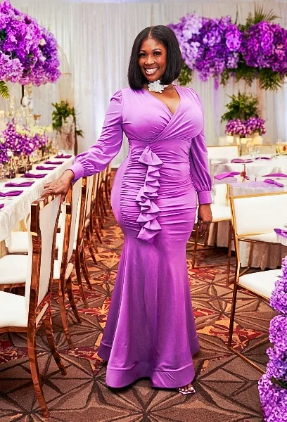 A woman in a purple dress standing in a banquet hall decorated with purple floral arrangements and white table settings.